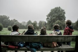 Children sitting on a wooden bench outside at a park. They are wearing coats and facing away from the camera. There are backpacks with them on the bench.