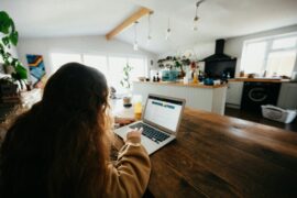 A teenahe girl is sitting with her back to the camera while looking at the screen of a laptop. She has long brown hair and is sitting at a wooden table in a dining room.