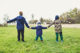 Three children outside, wearing winter coats and hats. All three are facing away from the camera so we only see their backs. They are holding hands. There are two taller children on either side and a younger, shorter child is in the middle.