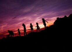 Photo shows a sunset with several children running down a gently sloping hill; one in front of the other. The sky is purple and orange, while the hill is in the shadow and appears black.