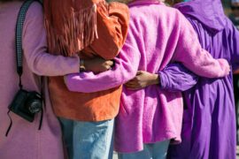 Community is necessary for the prevention of child sex trafficking. Here are four women united in community, walking arm-in-arm away from the camera. All four women are wearing jackets in shades of pink, orange, and purple.