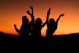 A photo showing human connection. Three people are shown celebrating or dancing at sunset. Their bodies are shown as sihlouettes against a bright orange sky. Their arms are waving in the air.