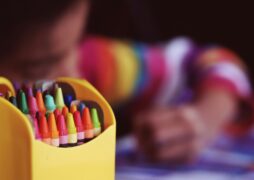 One way to practice mindfulness and relaxation is to encourage coloring. The child in this photo is using his left hand to color a picture. He is blurred in the background, while a yellow box of colorful crayons is featured prominantly in the lower left corner of the photo.