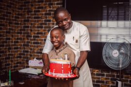 Fostering a positive mindset includes celebrating wins with your children like this father who is celebrating with his son by baking and decorating a cake in the shape of a red drum. They are standing in front of a window, a brick wall and a standing fan.