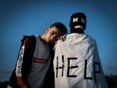 It is crucial for our children to know that they can ask for help. The young man in this photo is standing by another person whose back is to the camera. This person has a black knit hat on with white markings for eyes and a mouth. There is also a white garment with the word HELP in black written on it.