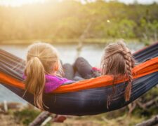 Helping children manage trauma triggers includes creating safe environments and spaces to relaxy like these two girls relaxing in a hammock overlooking the serene lake and trees.