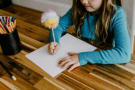 Allowing children to have a say in their routine and structure helps build their confidence. This child is sitting down at a table with a sheet of paper and a pencil with a pom-pom decoration on the end getting ready to write her plan for the week.