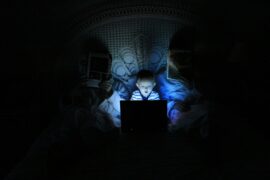 Implementing safe online practices is essential to protecting our children. This image shows two adults occupied with their own reading, blinding themselves to what the child in between them is viewing on the computer. All three are sitting in a bed in a very dark room illuminated only by the screen of the child's laptop.