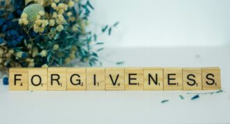 This photo shows the word forgiveness spelled out using Scrabble tiles. There is a bouquet of flowers behind the tiles.