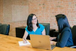 Having a trusted mentor can be invaluable to your growth. This image shows one woman mentoring another; two women sitting at a table. The woman on the right is speaking and the woman on the left is taking notes.